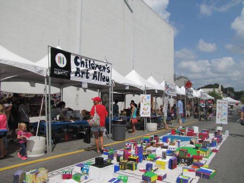 A vibrant street fair scene with tents labeled "Children's Art Alley," showcasing colorful art displays and visitors.