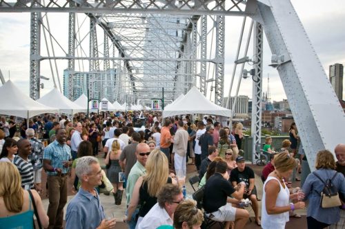 A crowded outdoor event on a bridge, with people socializing and white tents set up in the background.
