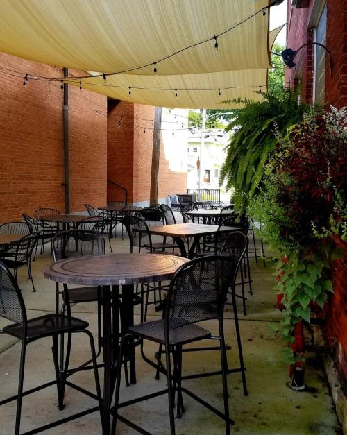 Outdoor dining area with empty metal tables, string lights, and hanging plants against a brick wall.