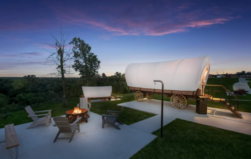 A scenic campsite with covered wagons, a fire pit, and benches, set against a colorful sunset sky.