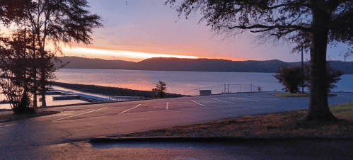 Sunset over a calm lake, with silhouettes of trees and a parking area in the foreground.