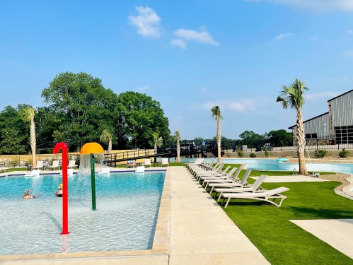 A sunny pool area with colorful water features, lounge chairs, and palm trees, surrounded by green grass and blue skies.