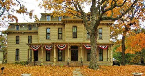 A large, historic house surrounded by colorful autumn leaves and decorated with red, white, and blue bunting.