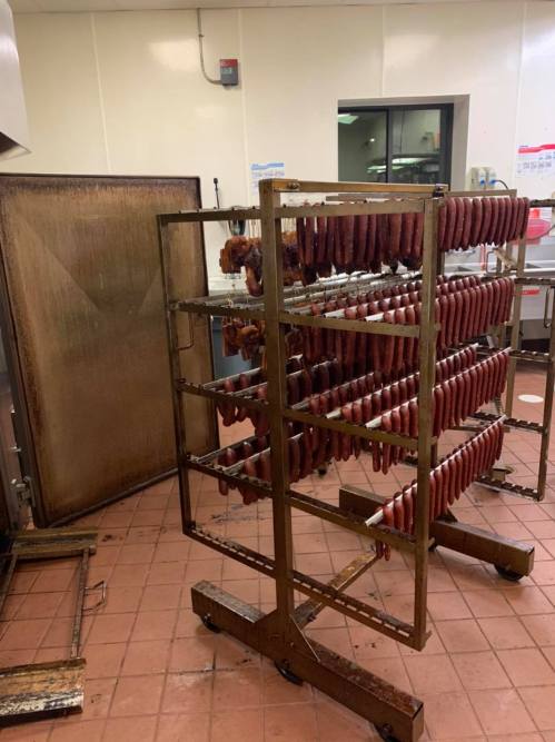 A metal rack filled with hanging sausages in a commercial kitchen, with a large oven in the background.