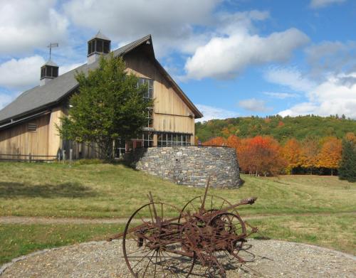 A rustic barn with a stone wall, surrounded by colorful autumn trees and a vintage plow in the foreground.