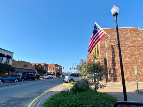 A view of a downtown street with an American flag, shops, and clear blue skies.