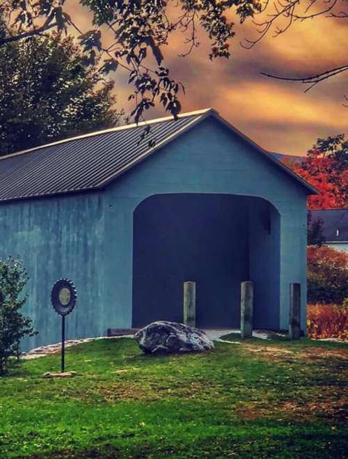 A blue shed with an open front, surrounded by greenery and a large rock, under a colorful sunset sky.
