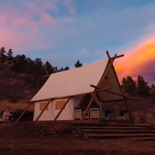 A canvas tent cabin on a hillside at sunset, with colorful clouds and trees in the background.