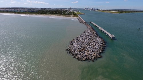 Aerial view of a rocky jetty extending into calm waters, with a coastal town and industrial area in the background.