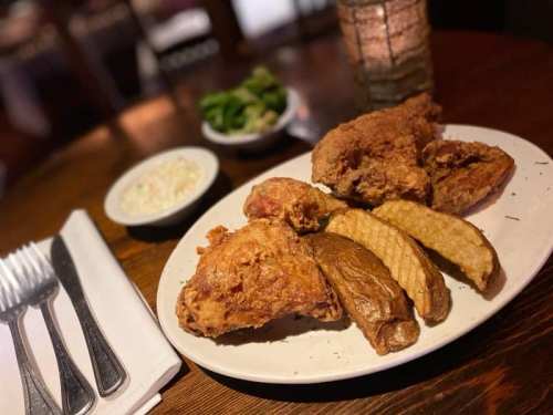 A plate of crispy fried chicken with sides of cornbread and coleslaw, set on a wooden table with utensils nearby.