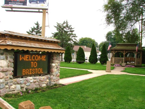 A stone sign reads "Welcome to Bristol" in a park with trees, flowers, and a gazebo in the background.
