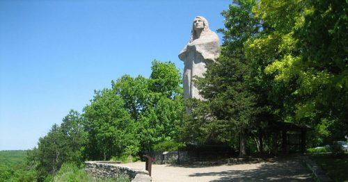 A large statue stands among trees on a sunny day, overlooking a winding path and lush green landscape.