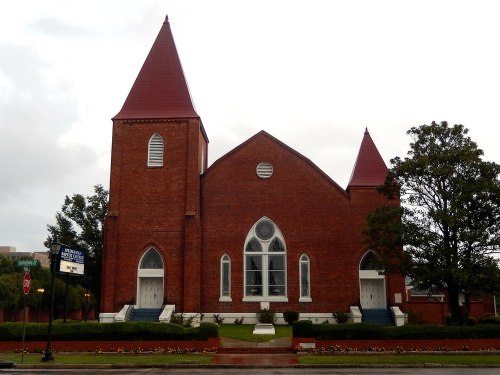 A red-brick church with two spires, large windows, and a landscaped front yard on a cloudy day.