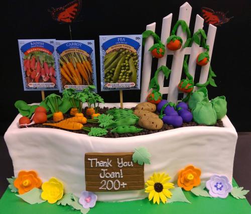 A cake designed as a garden bed with vegetables, seed packets, and a sign reading "Thank You Joan! 200+."