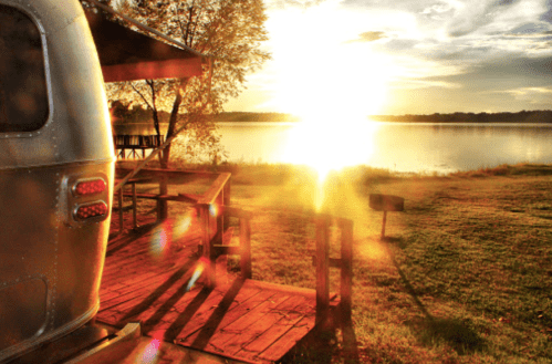 A serene lakeside view at sunset, featuring an Airstream trailer and golden light reflecting on the water.