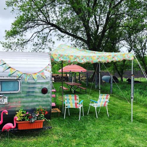 A vintage silver trailer with colorful decorations, surrounded by green grass, chairs, and pink flamingos.