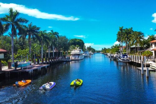 A scenic canal lined with palm trees, featuring boats and yachts under a bright blue sky.