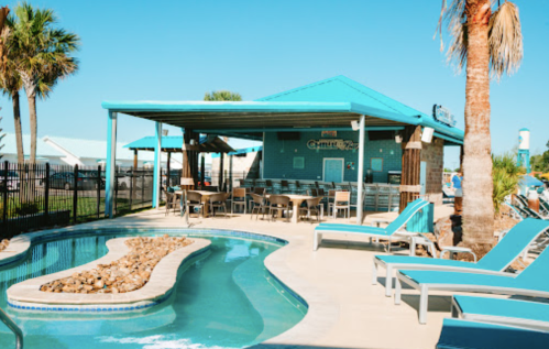 A sunny pool area with lounge chairs, a winding pool, and a blue-roofed building in the background. Palm trees are visible.