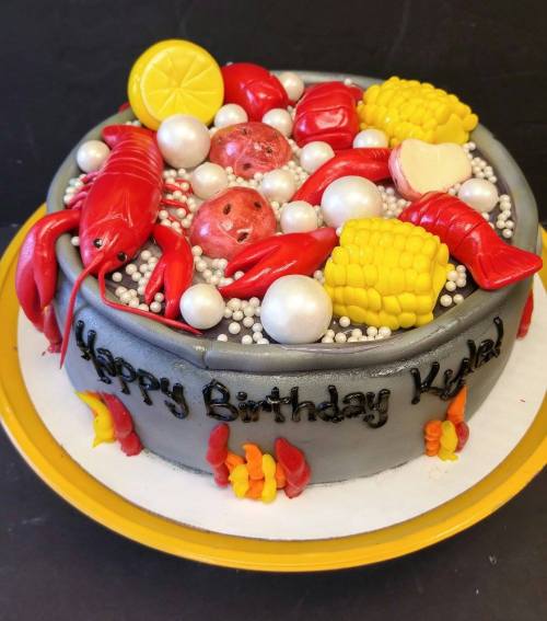 A birthday cake decorated with seafood, including lobsters, corn, and pearls, with "Happy Birthday Kadeal" written on it.