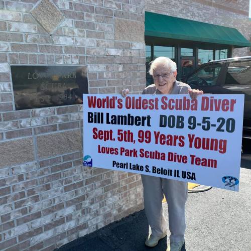 An elderly man holds a sign celebrating him as the world's oldest scuba diver, with details about his age and dive team.