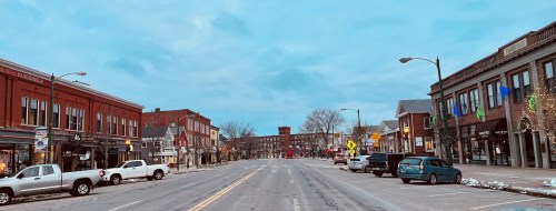 A quiet downtown street lined with shops and parked cars, under a cloudy sky. Snow is visible on the ground.
