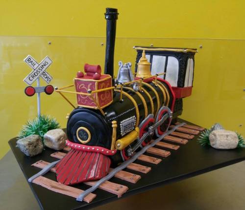 A colorful cake shaped like a vintage steam train on a track, with decorative rocks and a crossing sign.
