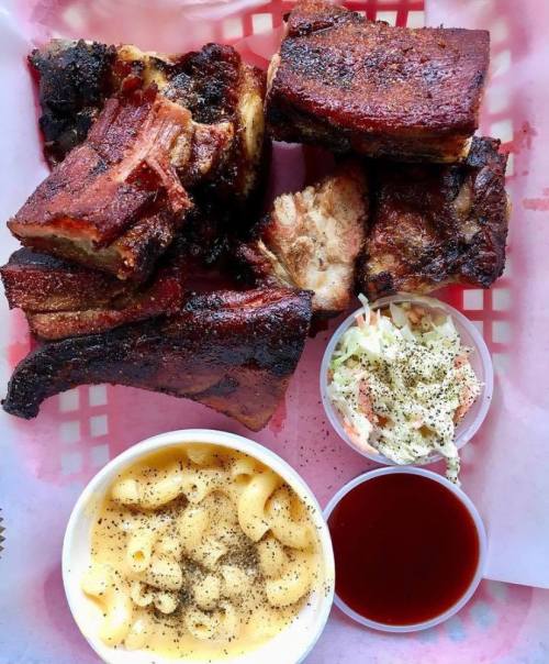 A platter of smoked ribs with coleslaw, macaroni and cheese, and barbecue sauce in a red-checkered basket.