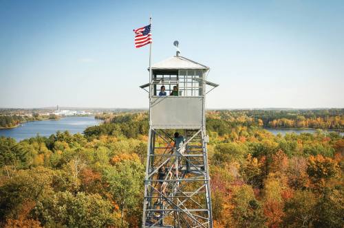 A tall fire tower with an American flag, surrounded by vibrant autumn trees and a scenic lake in the background.