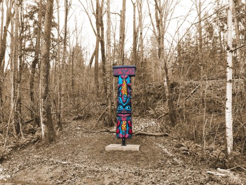 A colorful totem pole stands in a forested area, surrounded by trees and ferns, with a muted background.