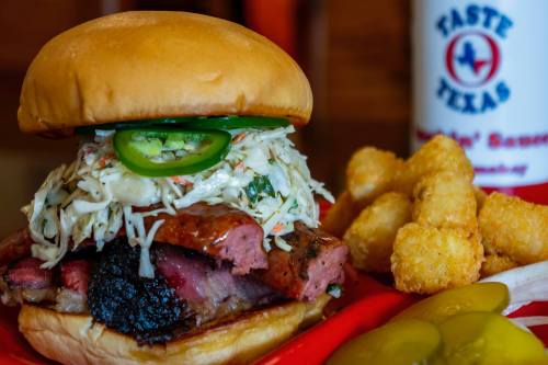 A close-up of a BBQ sandwich topped with coleslaw and jalapeños, served with tater tots and pickles on a red plate.