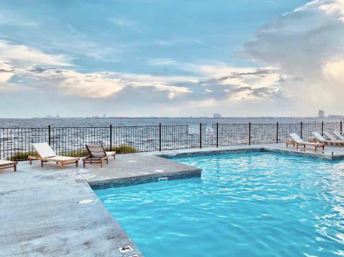 A serene poolside view with lounge chairs overlooking a calm body of water and a cloudy sky.