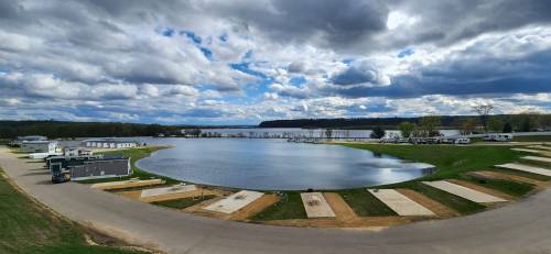 A scenic view of a lake surrounded by green grass and clouds, with RVs parked nearby and a road leading into the area.