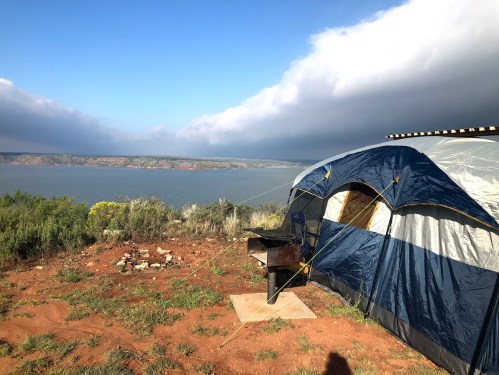 A blue and gray tent set up near a lake, with a scenic view of hills and cloudy skies in the background.
