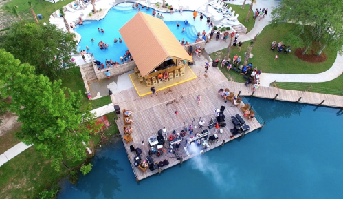 Aerial view of a lively pool area with a stage, people swimming, and a crowd enjoying a sunny day by the water.