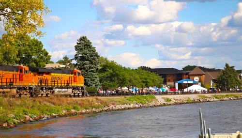 A train passes by a river with trees and a crowd at a festival in the background under a partly cloudy sky.