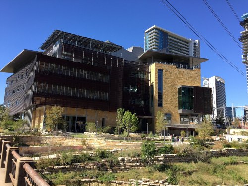 Modern building with a mix of stone and glass, surrounded by greenery and urban skyline in the background. Clear blue sky.