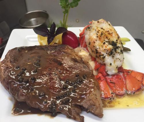 A gourmet plate featuring a grilled steak with sauce and a lobster tail, garnished with herbs and lemon.