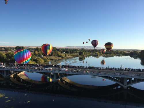 Colorful hot air balloons float over a river, with a crowd gathered on a bridge below, under a clear blue sky.