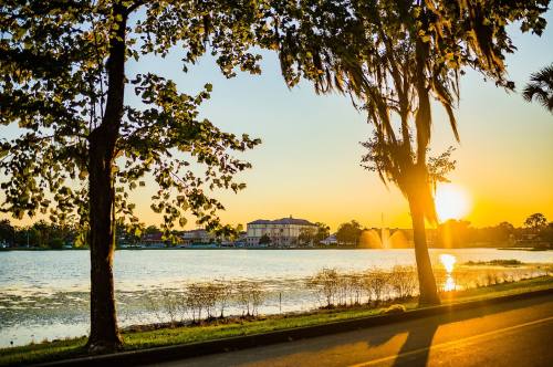 Sunset over a serene lake, framed by trees, with a building in the background and soft reflections on the water.