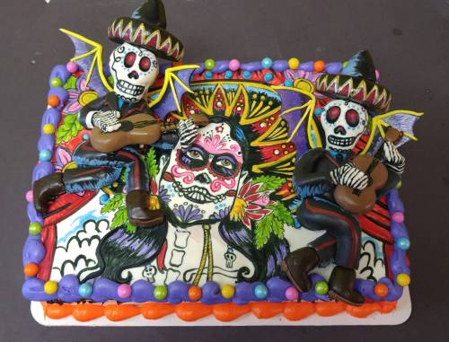 A colorful cake featuring skeleton figures in sombreros playing guitars, with a decorated sugar skull and vibrant designs.