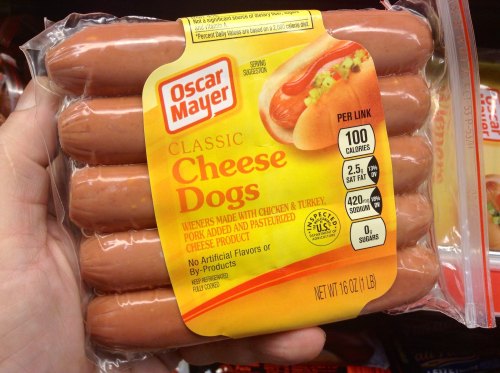 A hand holding a package of Oscar Mayer Classic Cheese Dogs, featuring six hot dogs and nutritional information on the label.