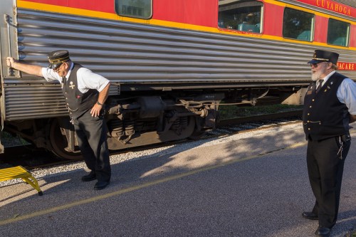 Two train conductors in uniforms stand beside a vintage train car, one leaning against it while the other observes.