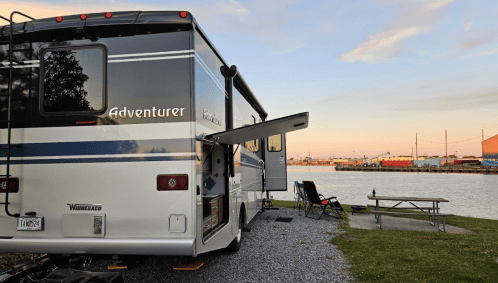 A parked RV by a calm waterway, with a picnic table and chairs nearby, under a colorful sunset sky.