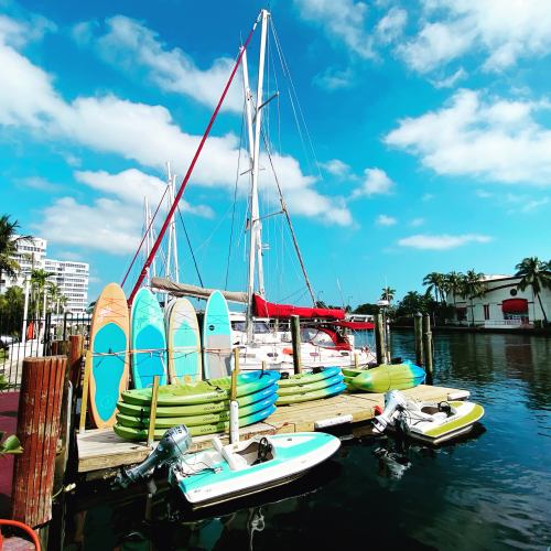 Colorful kayaks and paddleboards lined up on a dock beside a sailboat, with a clear blue sky and palm trees in the background.