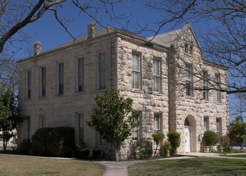 Historic stone building with large windows, surrounded by trees and shrubs, under a clear blue sky.