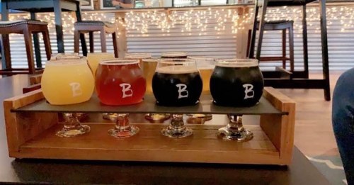 A wooden flight tray holds eight glasses of various craft beers, with a festive background of string lights.