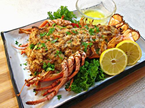 A baked lobster dish topped with breadcrumbs, garnished with parsley, lemon slices, and a side of melted butter.