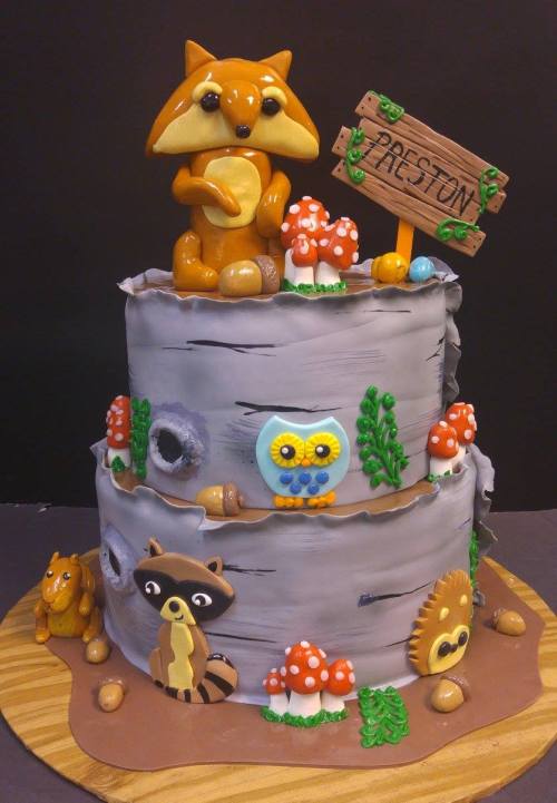 A whimsical two-tier cake decorated with woodland animals, mushrooms, and a sign reading "Preston."