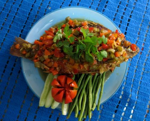 A whole fish topped with colorful diced vegetables, served with green beans and tomato slices on a blue plate.