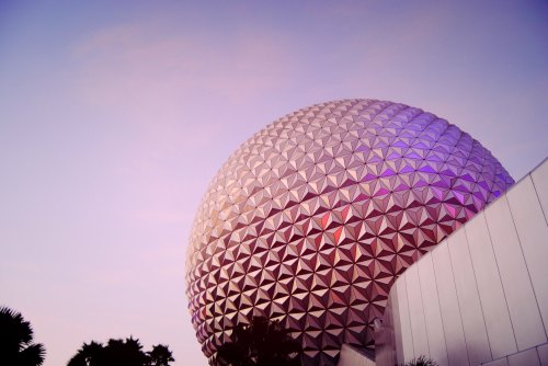 A large, geodesic dome structure against a pastel sky, with trees in the foreground and a modern building to the side.